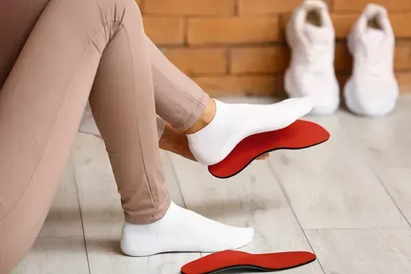 Persona que sostiene plantillas ortopédicas rojas, viste calcetines y mallas blancas y tiene zapatillas blancas de fondo.