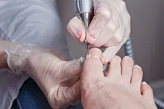 Persona que se está tratando la uña del pie con una herramienta rotatoria. Lleva guantes en las manos (primer plano).
