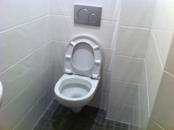 Petits toilettes avec WC et abattant surélevé dans un coin carrelé sous une plaque de chasse d'eau.