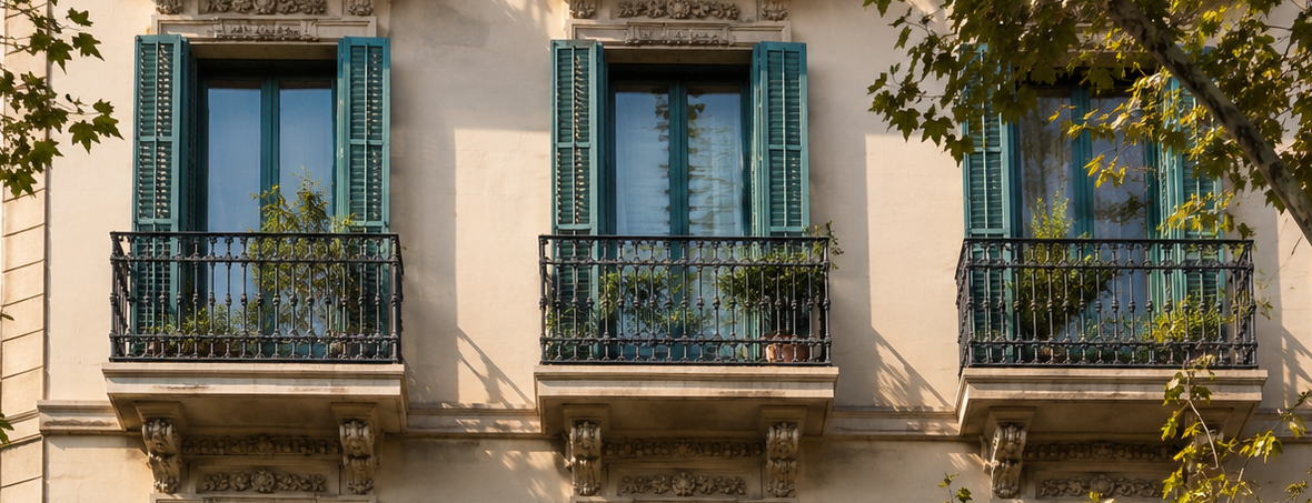 Una contraventana de madera está entreabierta en una ventana, dejando ver macetas con plantas en el alféizar y una vista borrosa de los edificios de la ciudad.