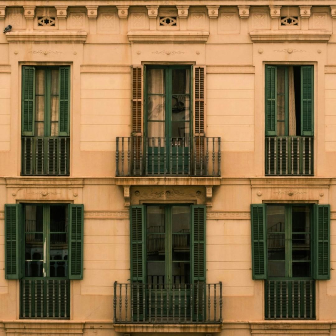 Un sofá beige con cojines rojos se encuentra frente a unas ventanas luminosas con contraventanas blancas, junto a una silla de comedor de madera.