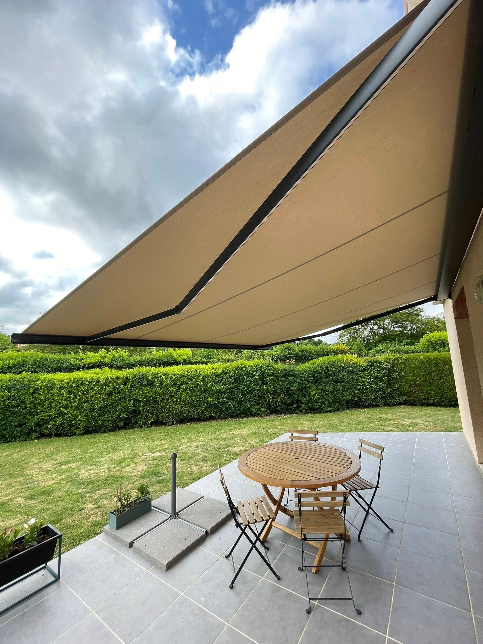 Un auvent beige protège une table et des chaises de patio en bois, sur une terrasse donnant sur une pelouse verte et une haie.