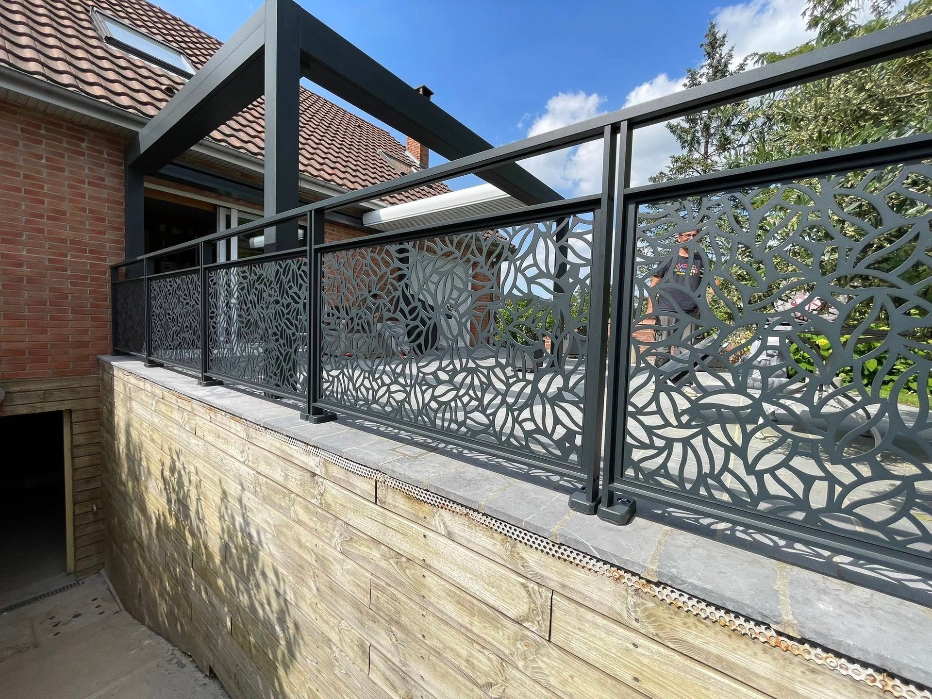 Clôture en métal noir découpé au laser, ornée de motifs de feuilles, surmontant un mur en pierre, à côté d'un bâtiment doté d'une pergola noire.