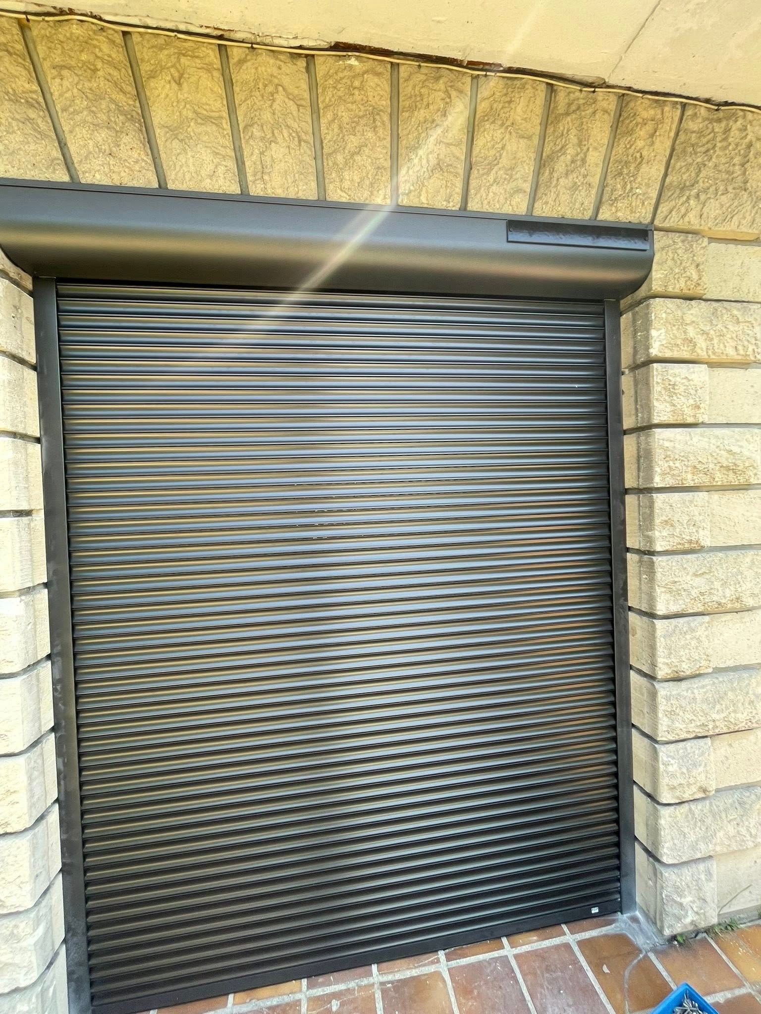 Un volet de sécurité noir est fermé sur une porte encadrée de briques beiges.