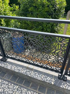 Rambarde de balcon en métal noir ornée de motifs de feuilles, projetant des ombres sur la terrasse en briques en contrebas. Feuillage vert en arrière-plan.