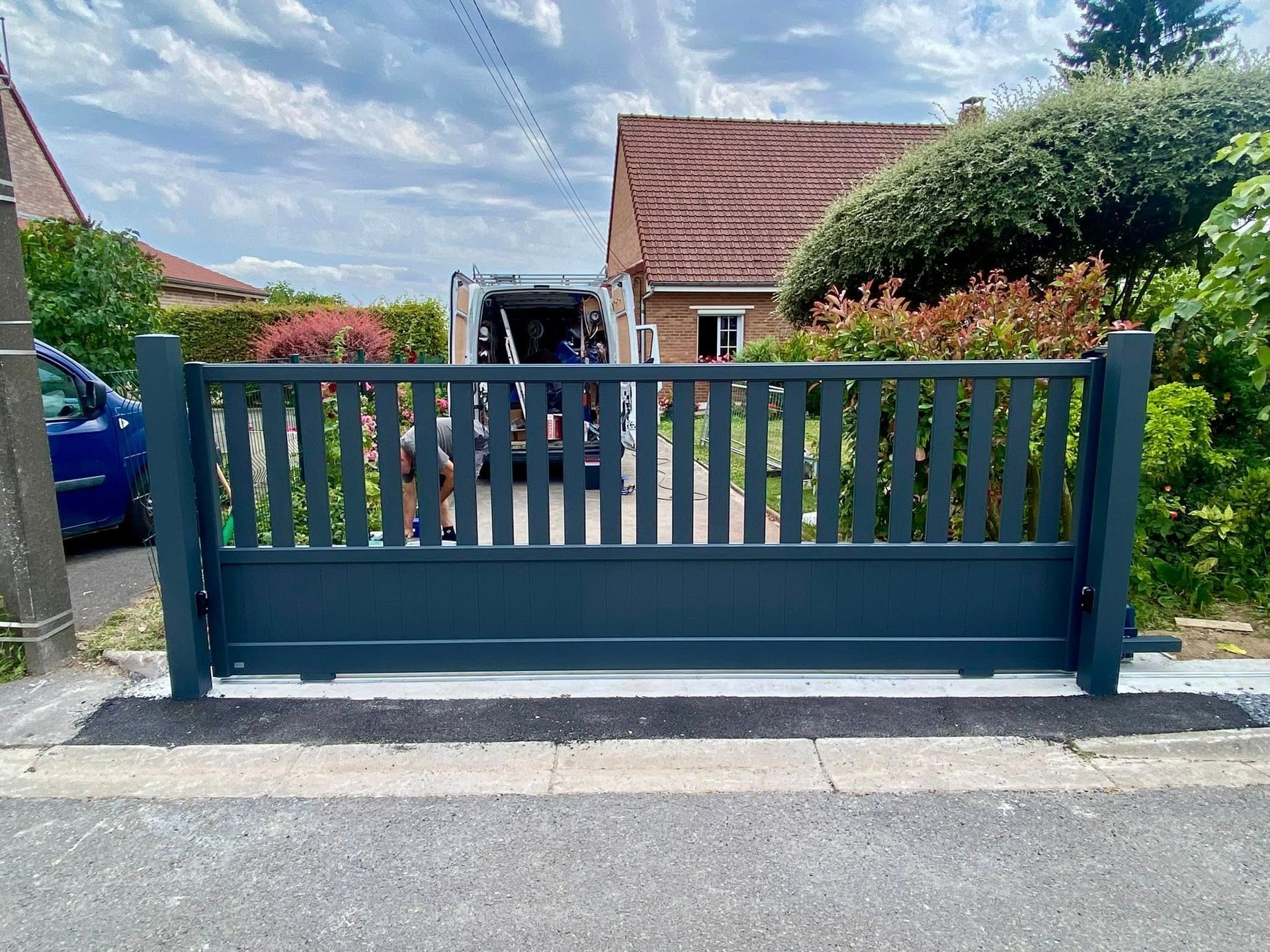 Portail coulissant bleu foncé devant une maison, posé sur du béton, avec une voiture en arrière-plan.