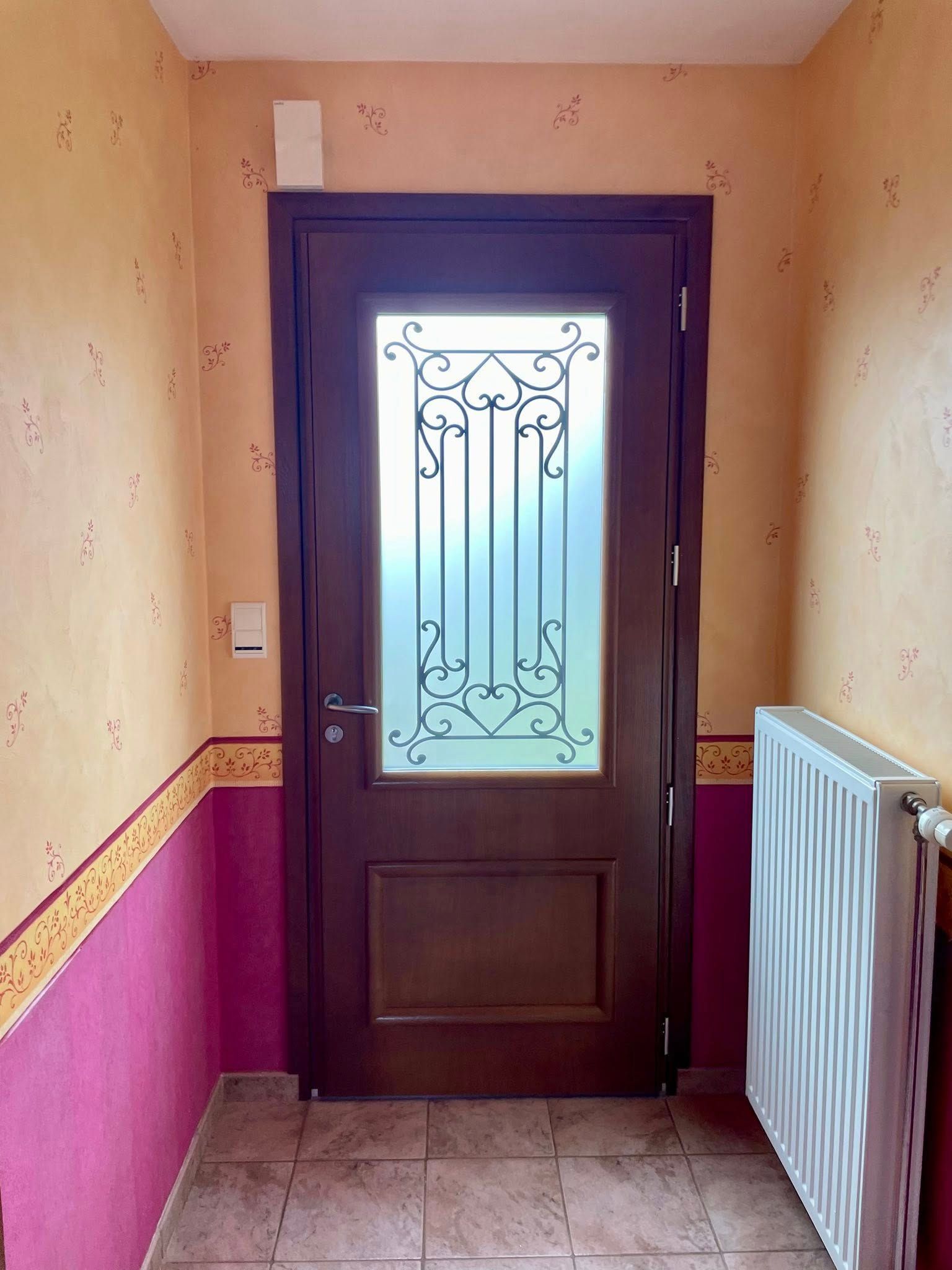 Porte marron avec panneau de verre et ornement métallique, dans un couloir aux murs roses et beiges, avec radiateur.