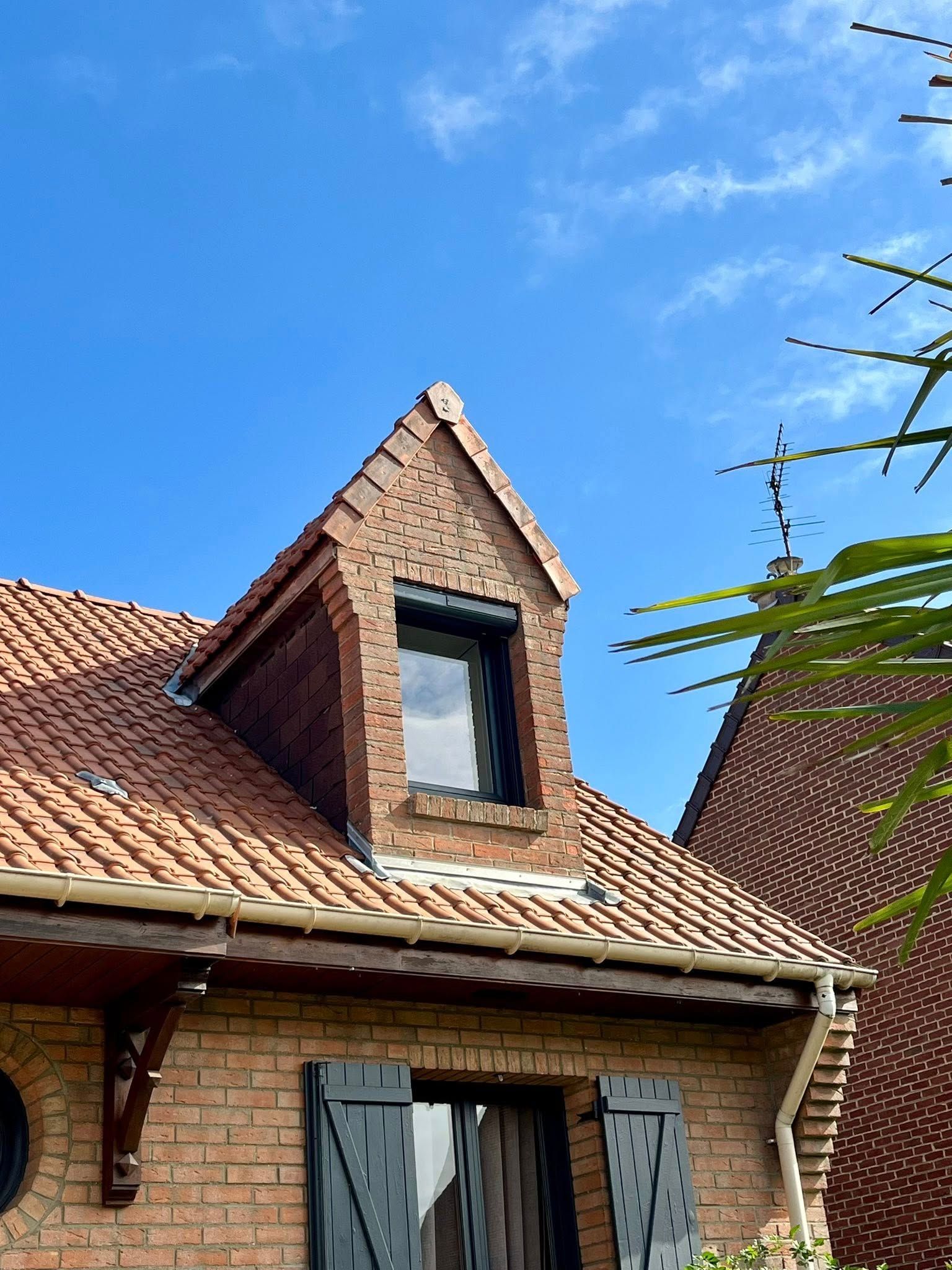 Maison en briques avec une lucarne, un toit de tuiles rouges et des volets sombres se détachant sur un ciel bleu.