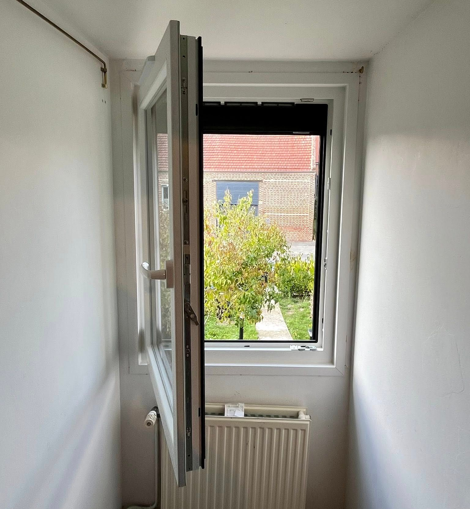 Fenêtre ouverte dans un espace étroit aux murs blancs, dévoilant de la verdure extérieure et un bâtiment en briques ; radiateur sous la fenêtre.