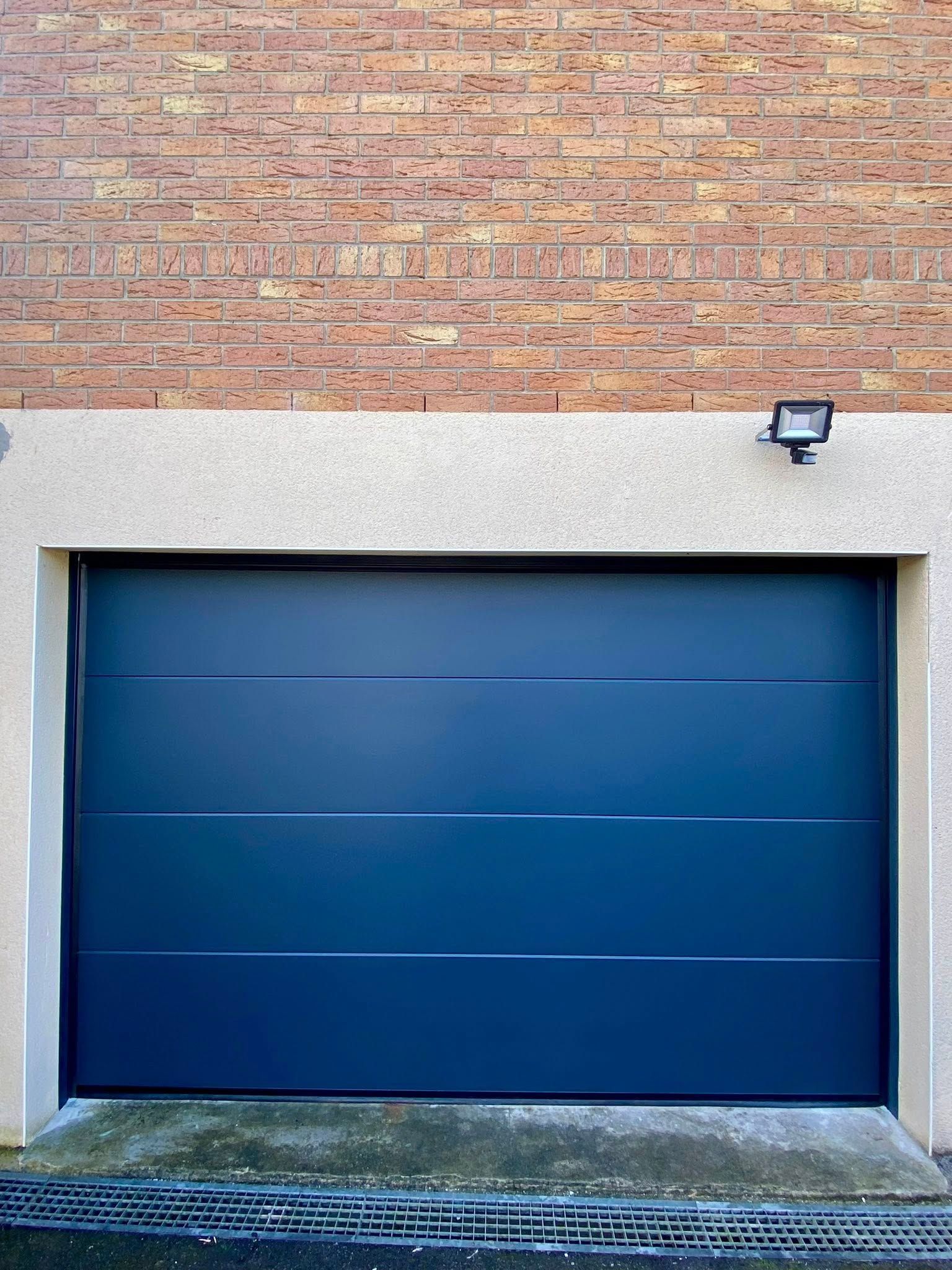 Porte de garage bleu foncé à rayures horizontales, sous un cadre en béton clair et un mur de briques.