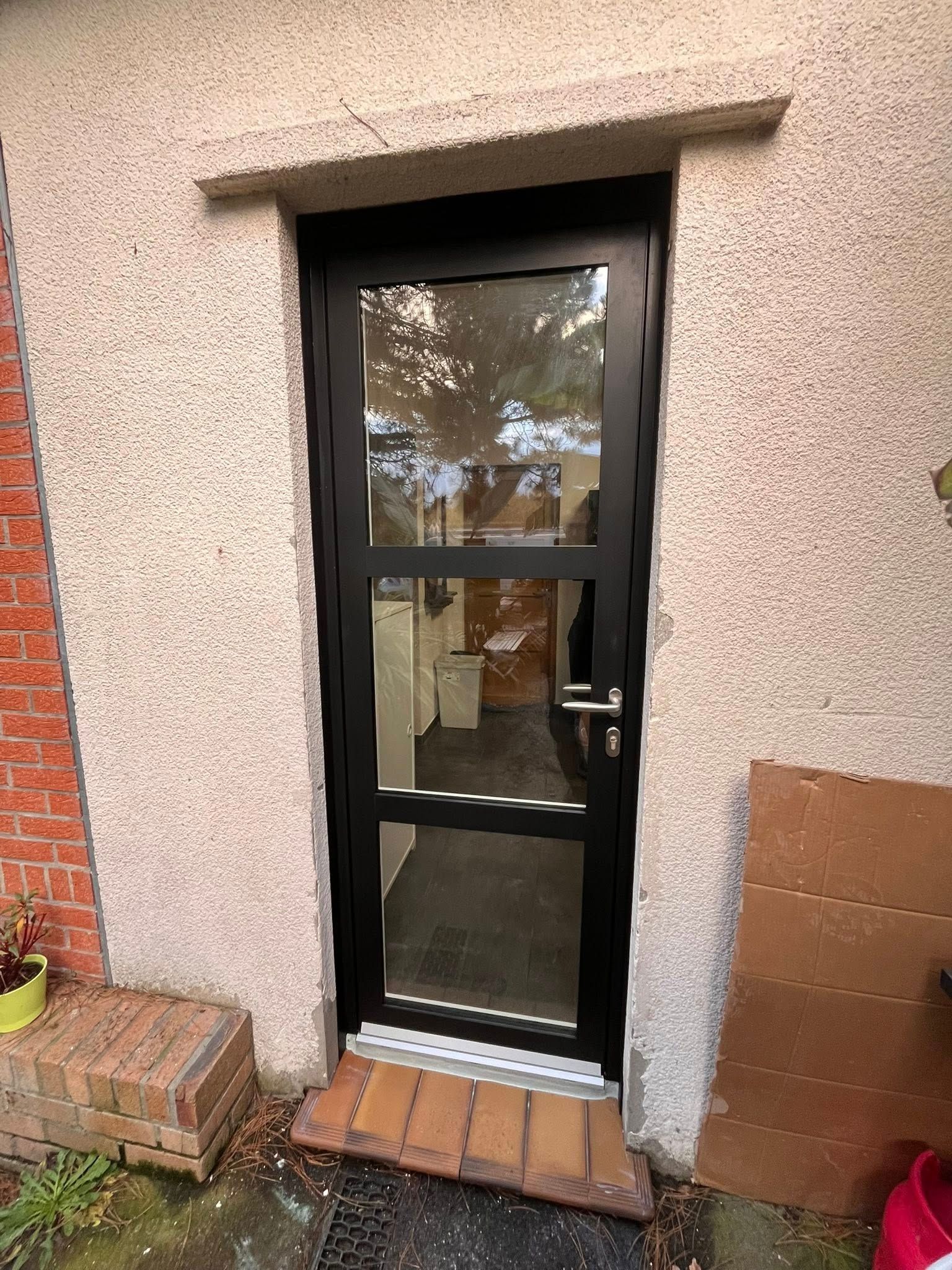 Porte vitrée à cadre noir sur un bâtiment en stuc. Un seuil en briques et un paillasson se trouvent à sa base.