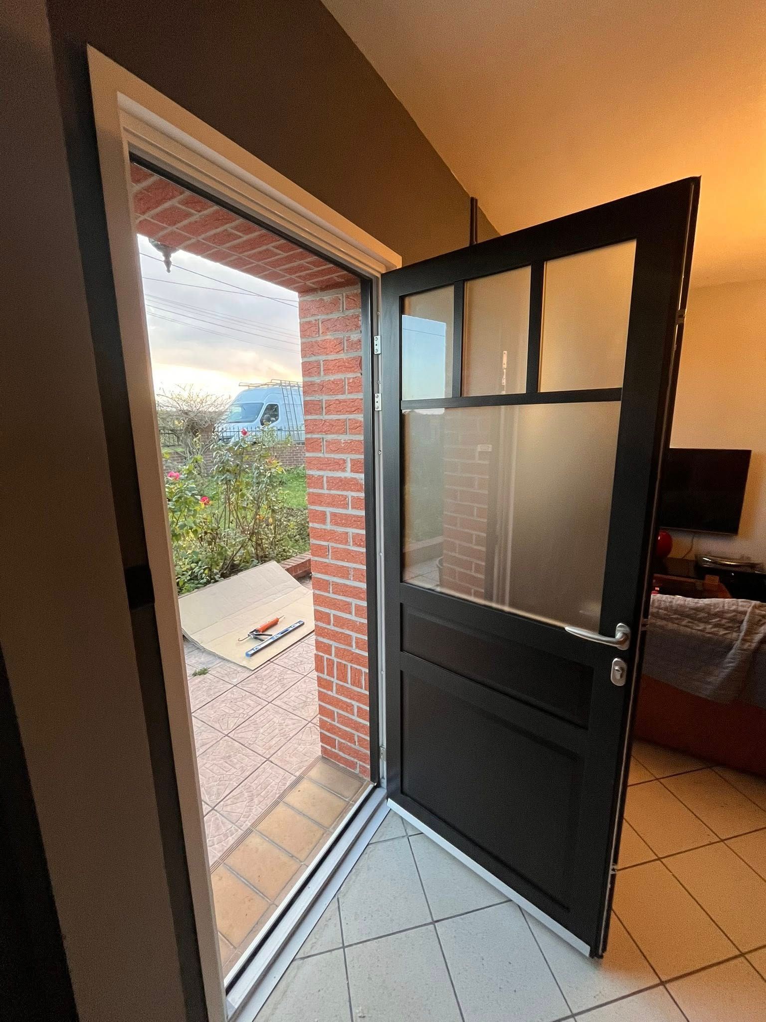 Porte noire vitrée ouverte, donnant sur un mur de briques extérieur.