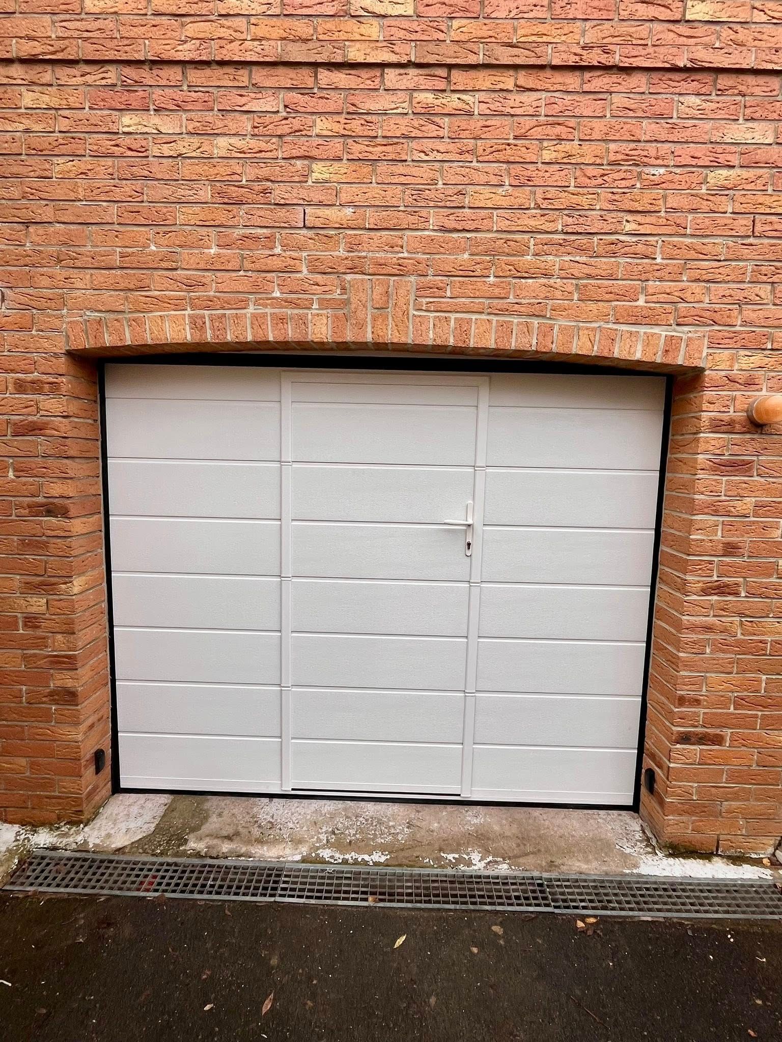 Porte de garage blanche à panneaux texturés, encastrée dans un mur de briques.