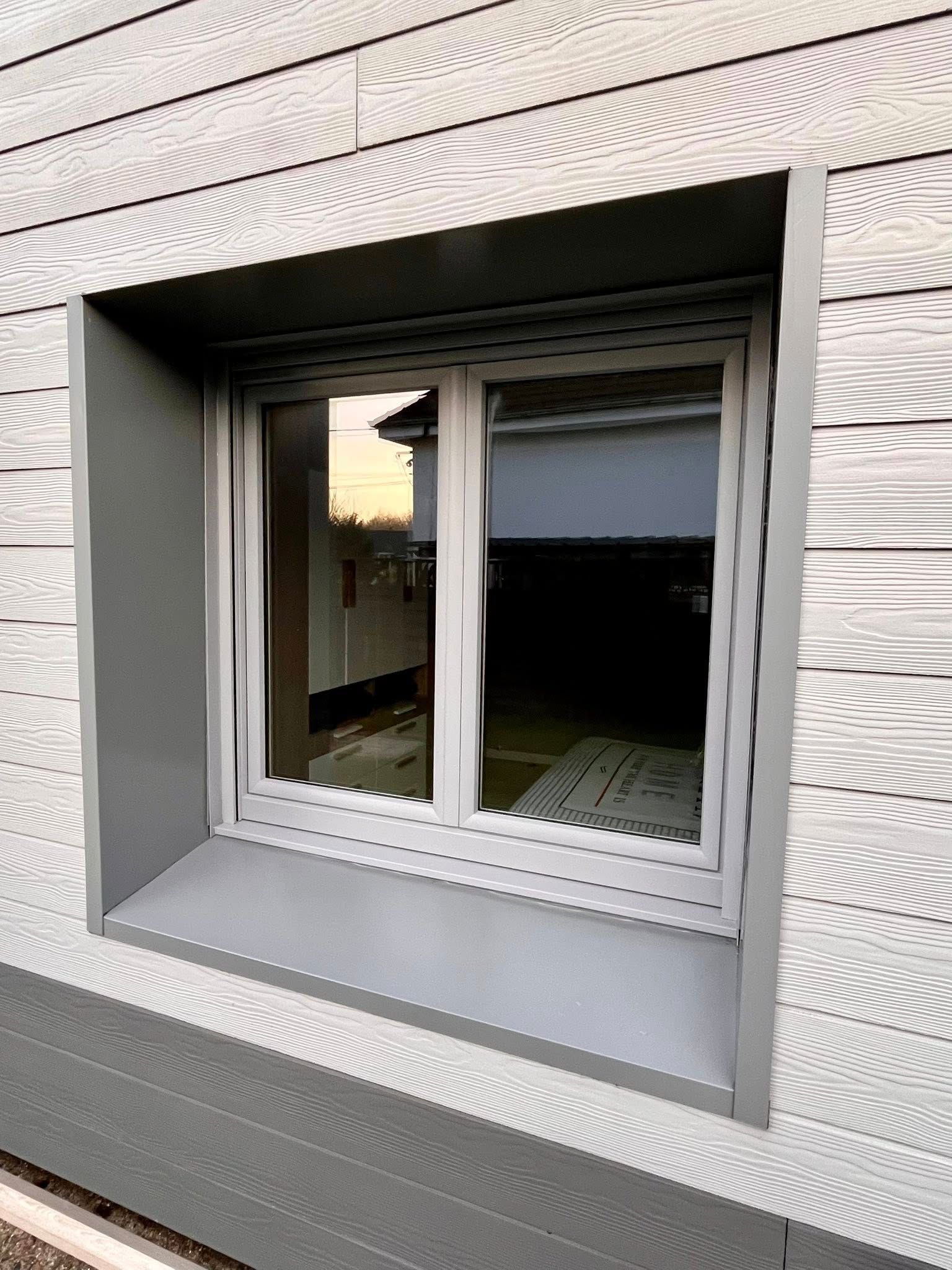 Fenêtre grise dans un cadre gris encastré dans un bardage. La fenêtre est équipée d'un store enrouleur et présente des reflets.
