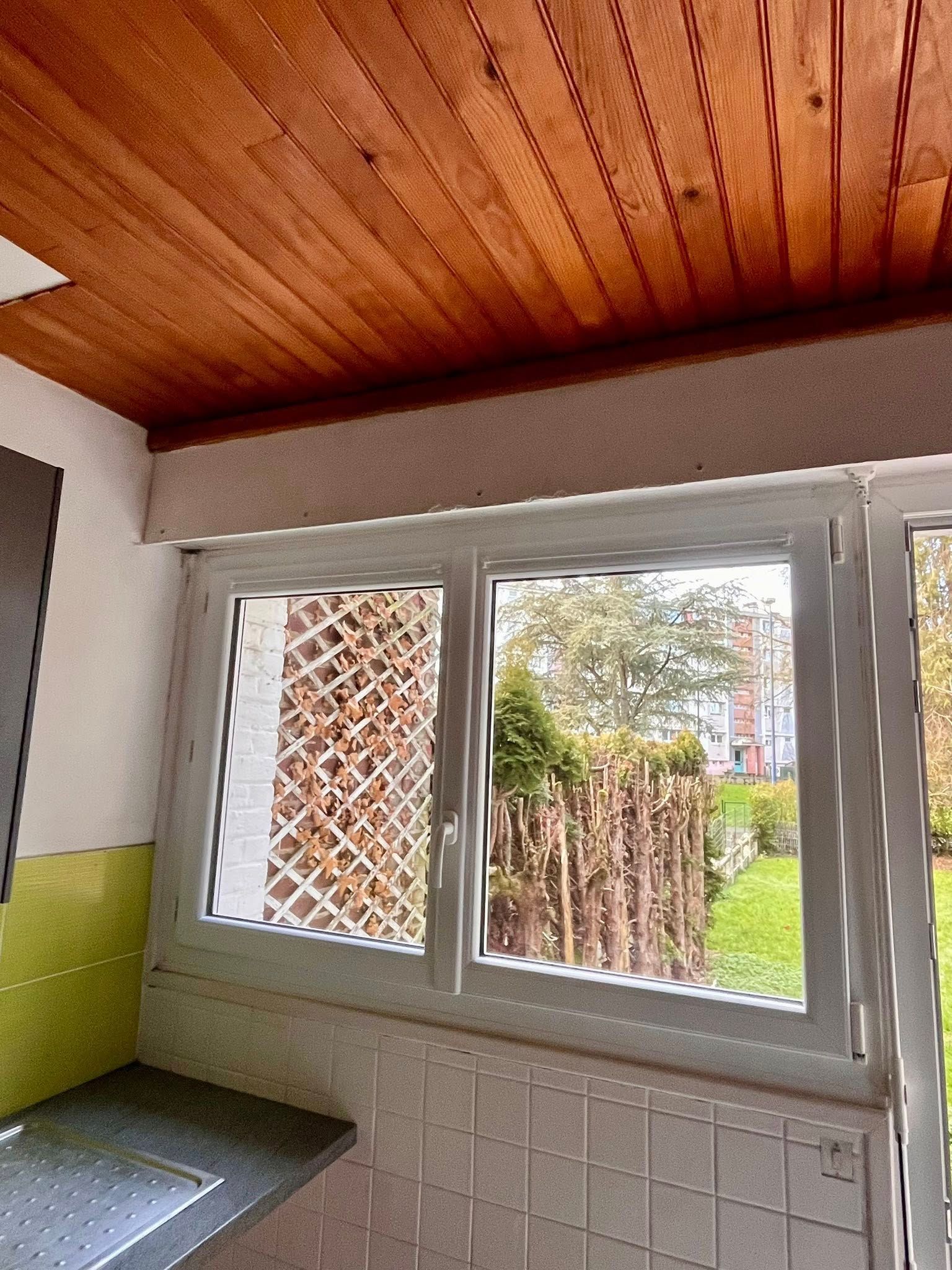 Fenêtre à cadre blanc dans une cuisine, donnant sur un jardin. Plafond en bois et carrelage blanc.