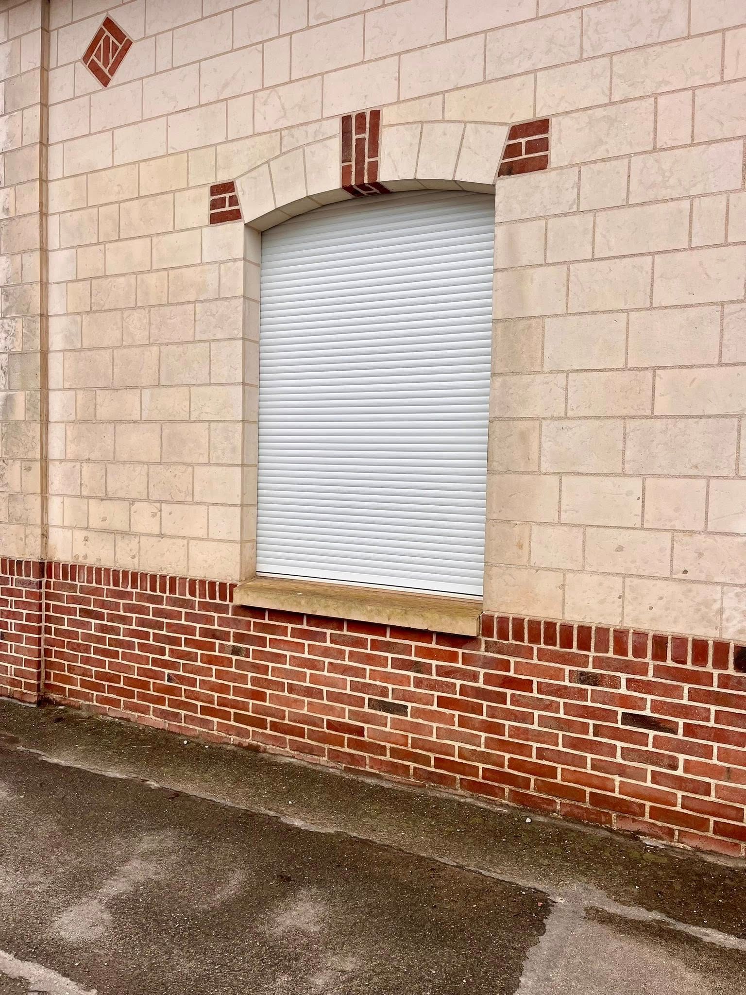 Fenêtre aux stores blancs fermés sur un bâtiment en briques avec un soubassement et des détails en briques rouges contrastantes.