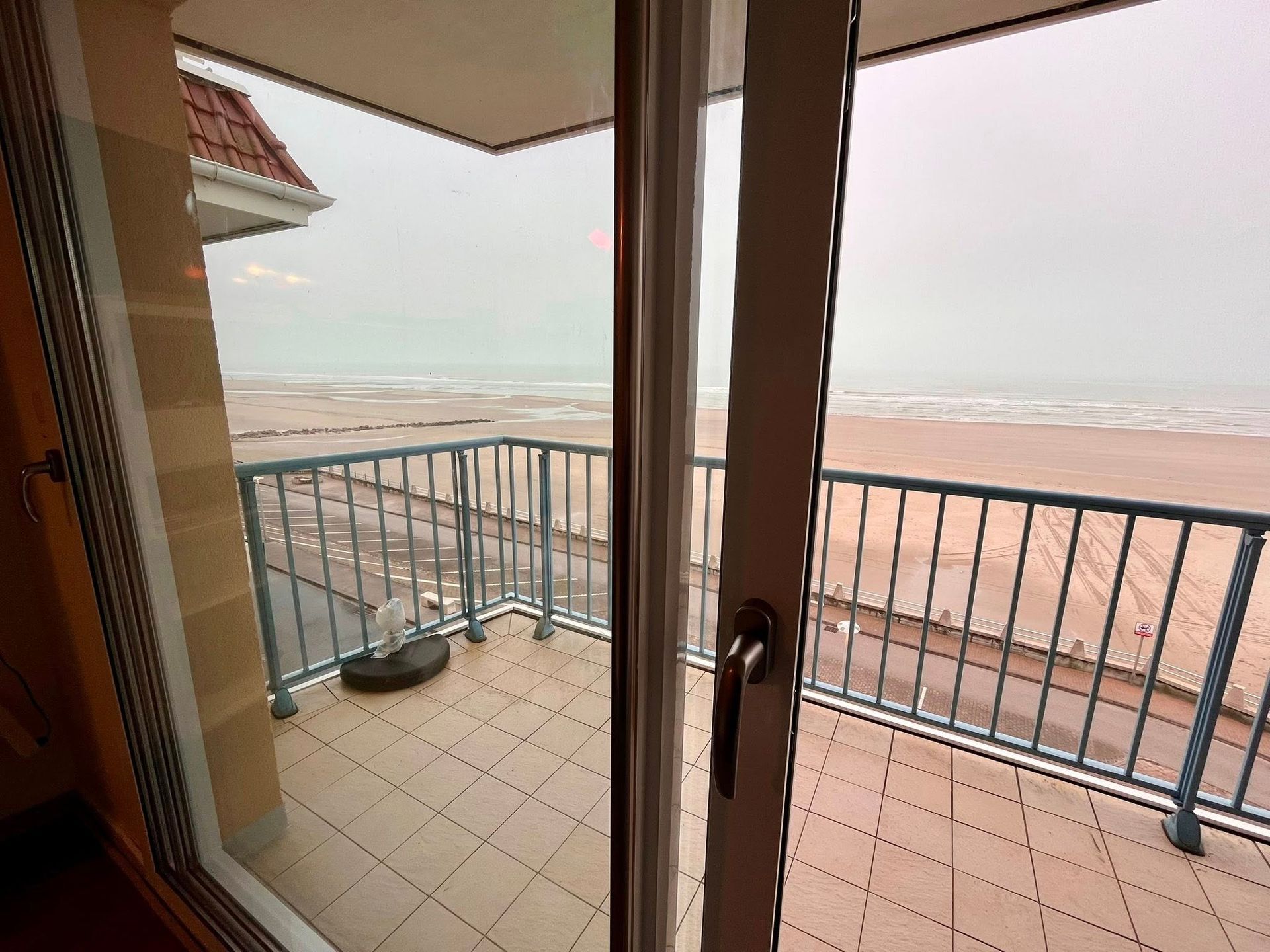 Vue sur la plage et l'océan depuis le balcon, à travers une porte-fenêtre coulissante. Ciel couvert, rambarde bleue, sol carrelé.