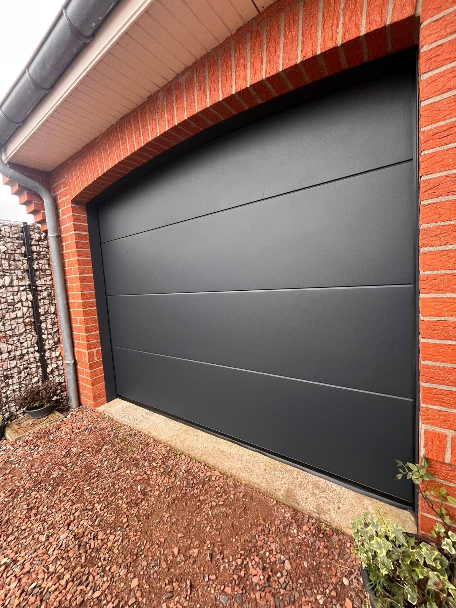 Porte de garage noire dans une arche en briques, sur un sol en gravier.