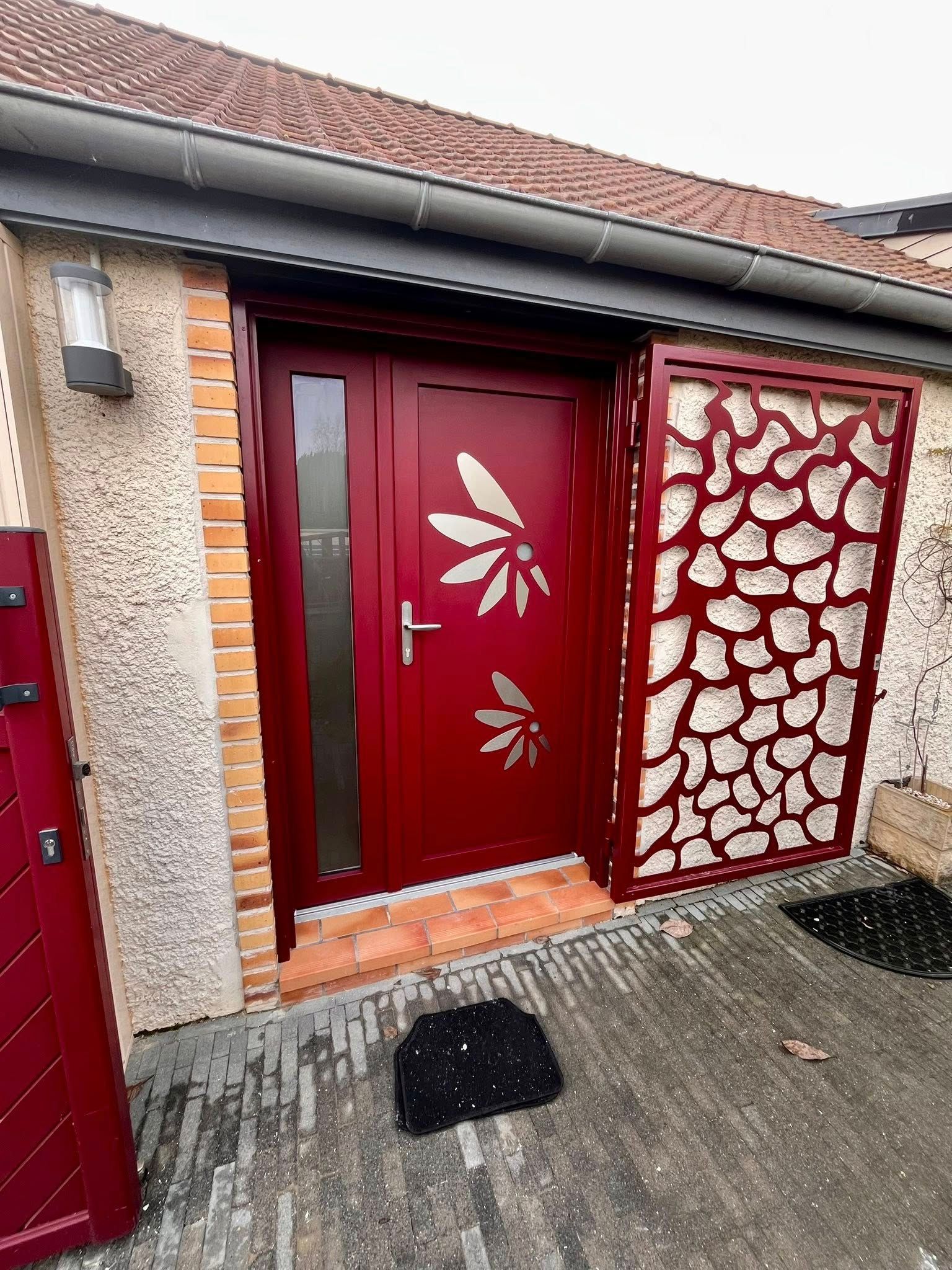 Porte d'entrée rouge ornée de motifs de feuilles, panneau latéral et paillasson. Façade en brique et en pierre.