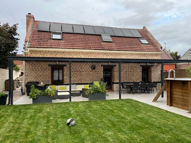 Jardin avec terrasse, pelouse et maison équipée de panneaux solaires. Pergola noire au-dessus du coin salon extérieur.