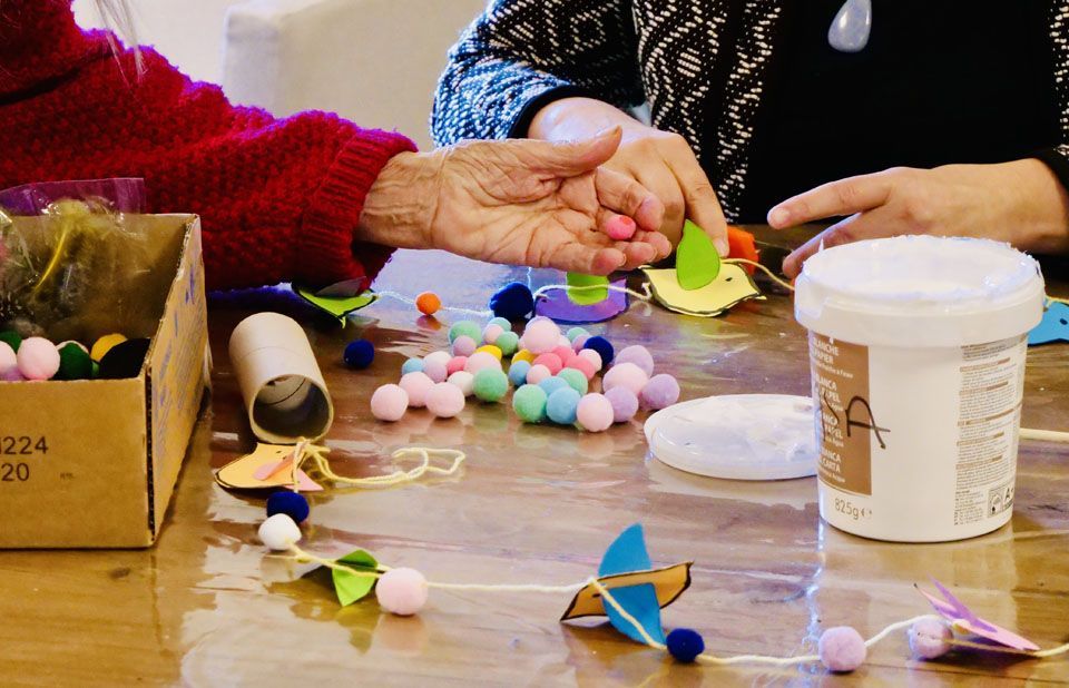 Des mains bricolent avec des pompons et des formes en papier sur une table en bois. Une boîte, de la colle et un tube en papier sont également présents.