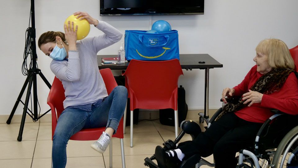 Une femme masquée lance une balle à une femme en fauteuil roulant ; il se passe quelque chose dans la pièce.