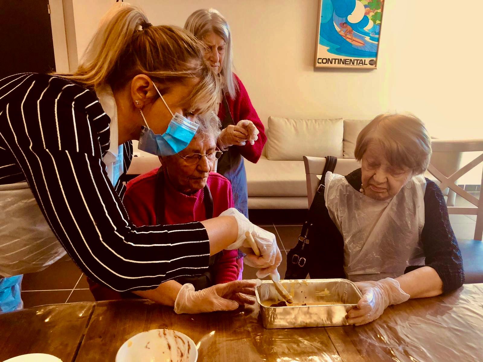 Une femme portant un masque aide deux femmes âgées à préparer de la nourriture à une table, une autre femme regarde dans un décor familial.