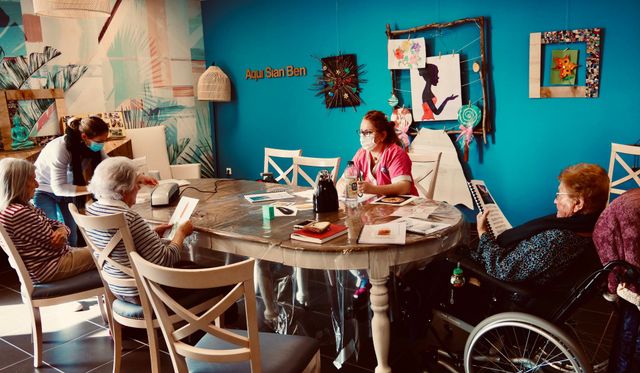 Des personnes âgées à une table bricolent avec un soignant dans une salle d'art colorée.