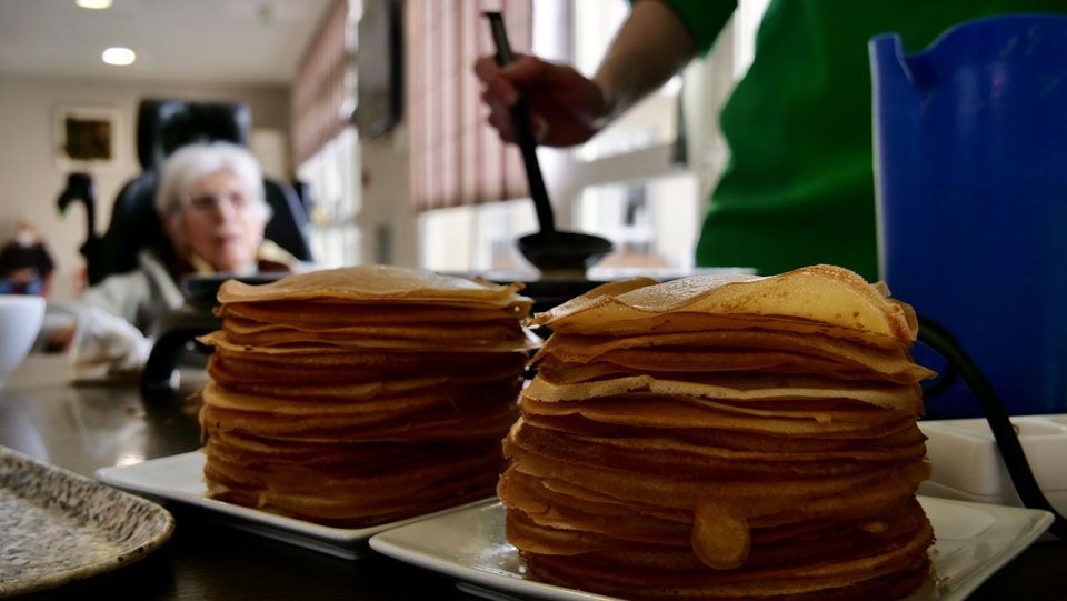 Deux piles de crêpes sur des assiettes blanches ; une personne en chemise verte remue ; une personne en fauteuil roulant regarde.