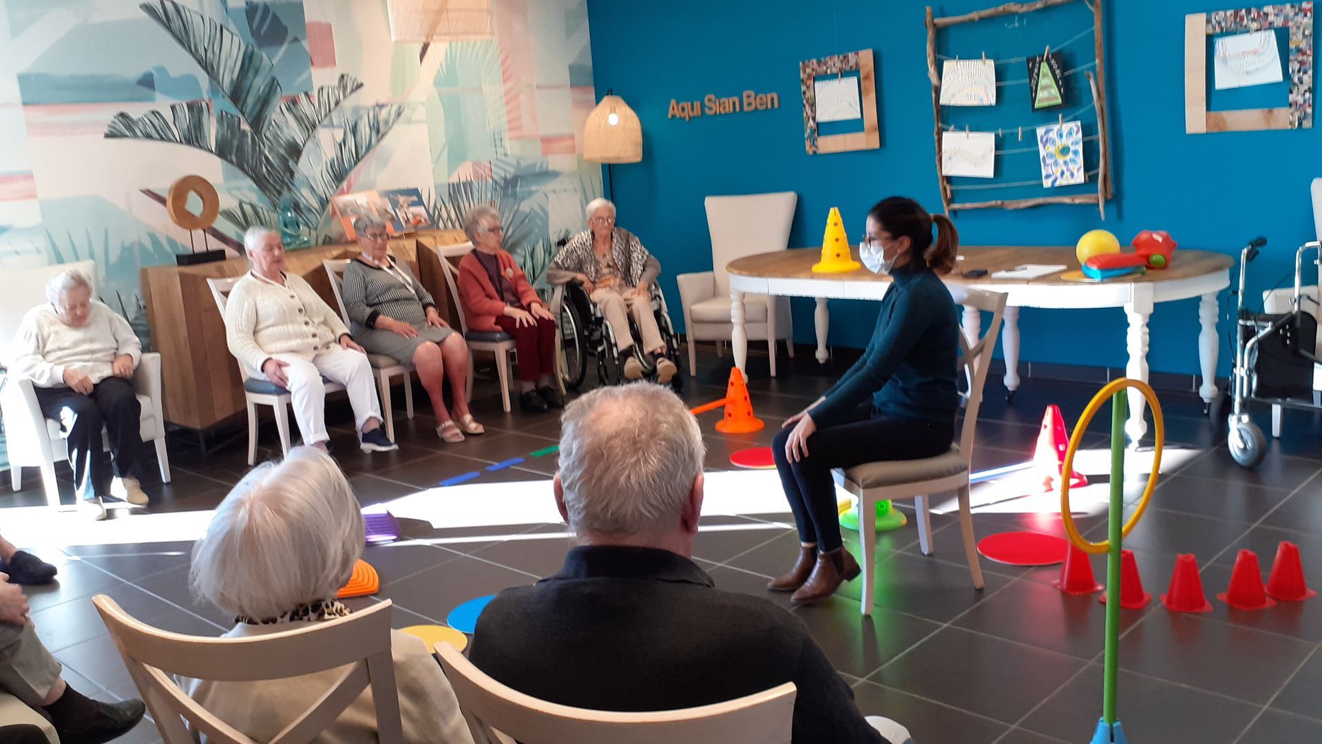 Cours d'exercices pour seniors : moniteur avec masque, assis ; seniors assis sur des chaises, matériel coloré, salle bleue.