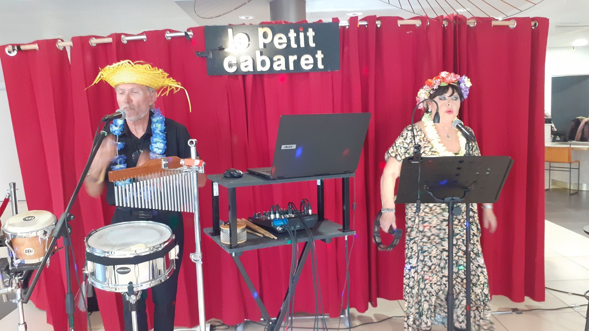 Deux artistes sur scène au « Petit Cabaret ». Un homme au chapeau de paille joue des instruments, une femme chante, avec un rideau rouge en arrière-plan.