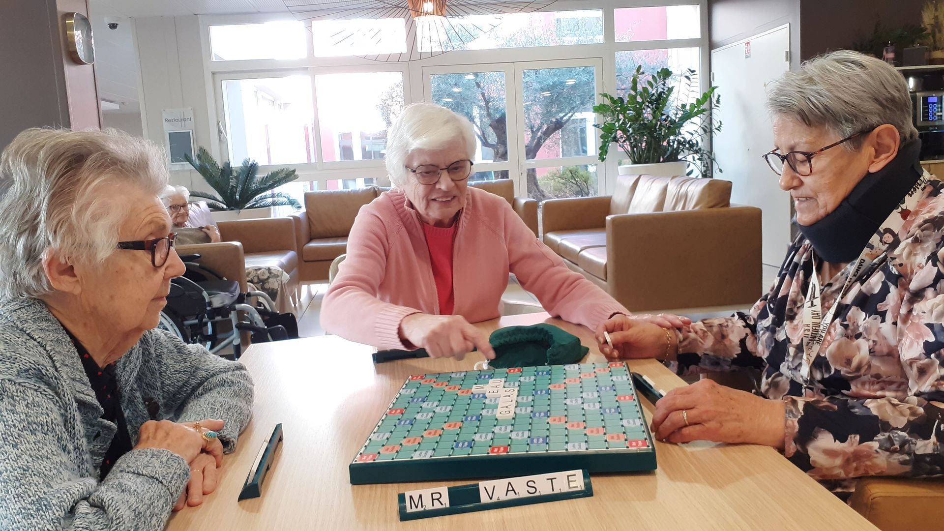 Trois femmes âgées jouent au Scrabble à une table dans un espace commun.