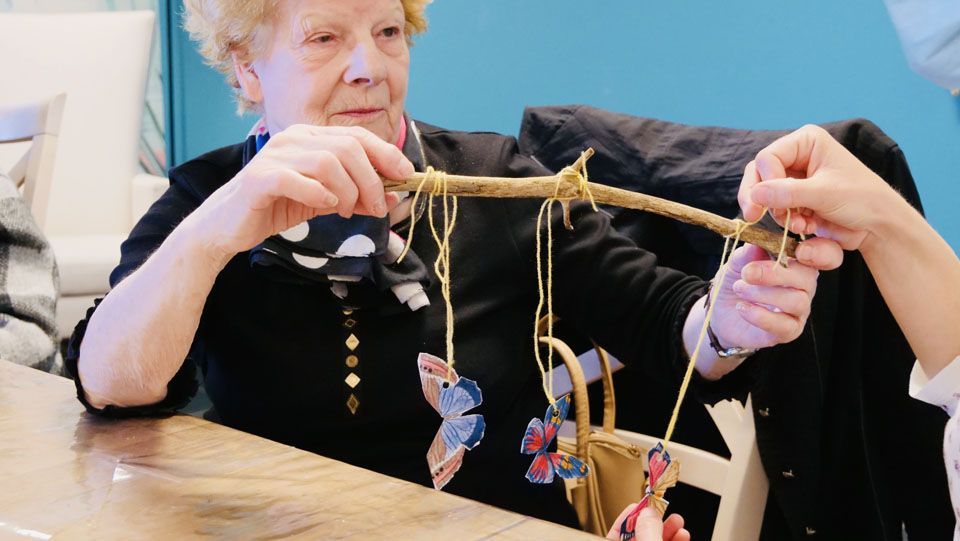 Une retraité réalise un objet décoratif. Elle tient un bâton auquel sont suspendus des papillons en papier. À l'intérieur.