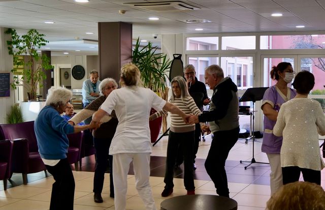 Des gens dansant en cercle dans une pièce bien éclairée, peut-être un centre pour personnes âgées ou un établissement de soins.