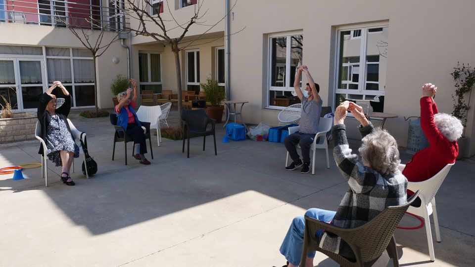 Un groupe de personnes fait de l'exercice assis sur des chaises à l'extérieur, les bras levés. Des bâtiments sont visibles en arrière-plan.