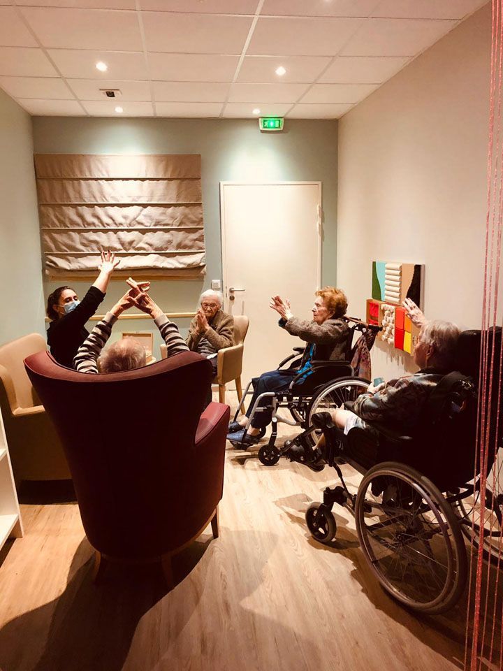 Groupe de personnes, certaines en fauteuil roulant, les bras levés, participant peut-être à une activité de groupe dans une pièce bien éclairée.