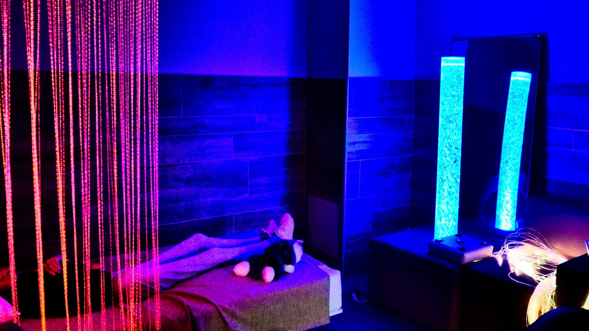 Pièce éclairée en bleu avec des colonnes décoratives, un rideau et une personne se reposant sur un lit avec une peluche.