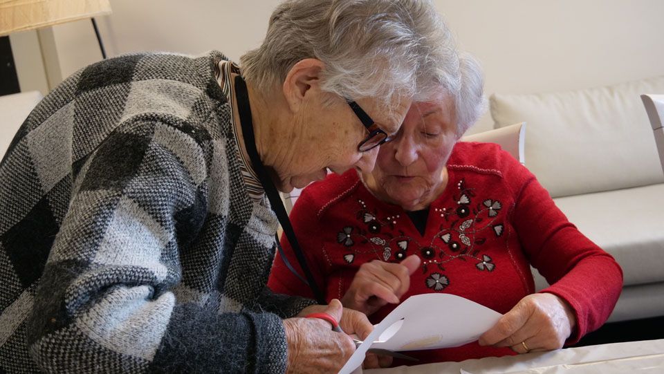 Deux femmes âgées, assises à l'intérieur, examinent attentivement un document. L'une d'elles montre du doigt en lisant.