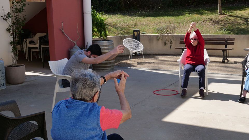 Exercices de groupe en extérieur. Des personnes assises sur des chaises étirent les bras. Journée ensoleillée.