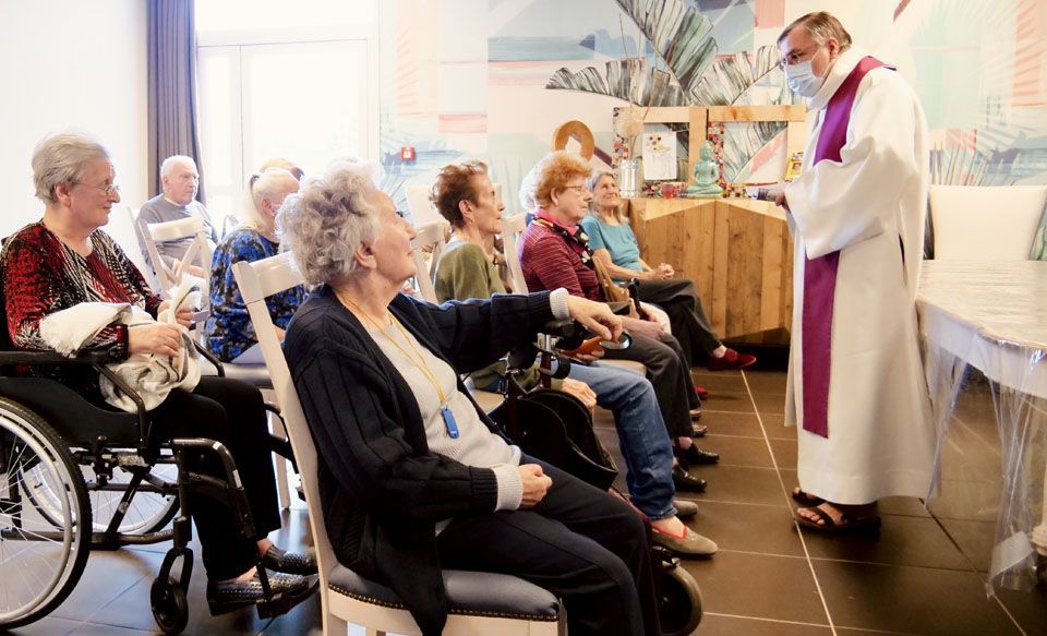 Un prêtre en habits liturgiques dirige un office pour les résidents de la maison de retraite dans une salle de l'ehpad.