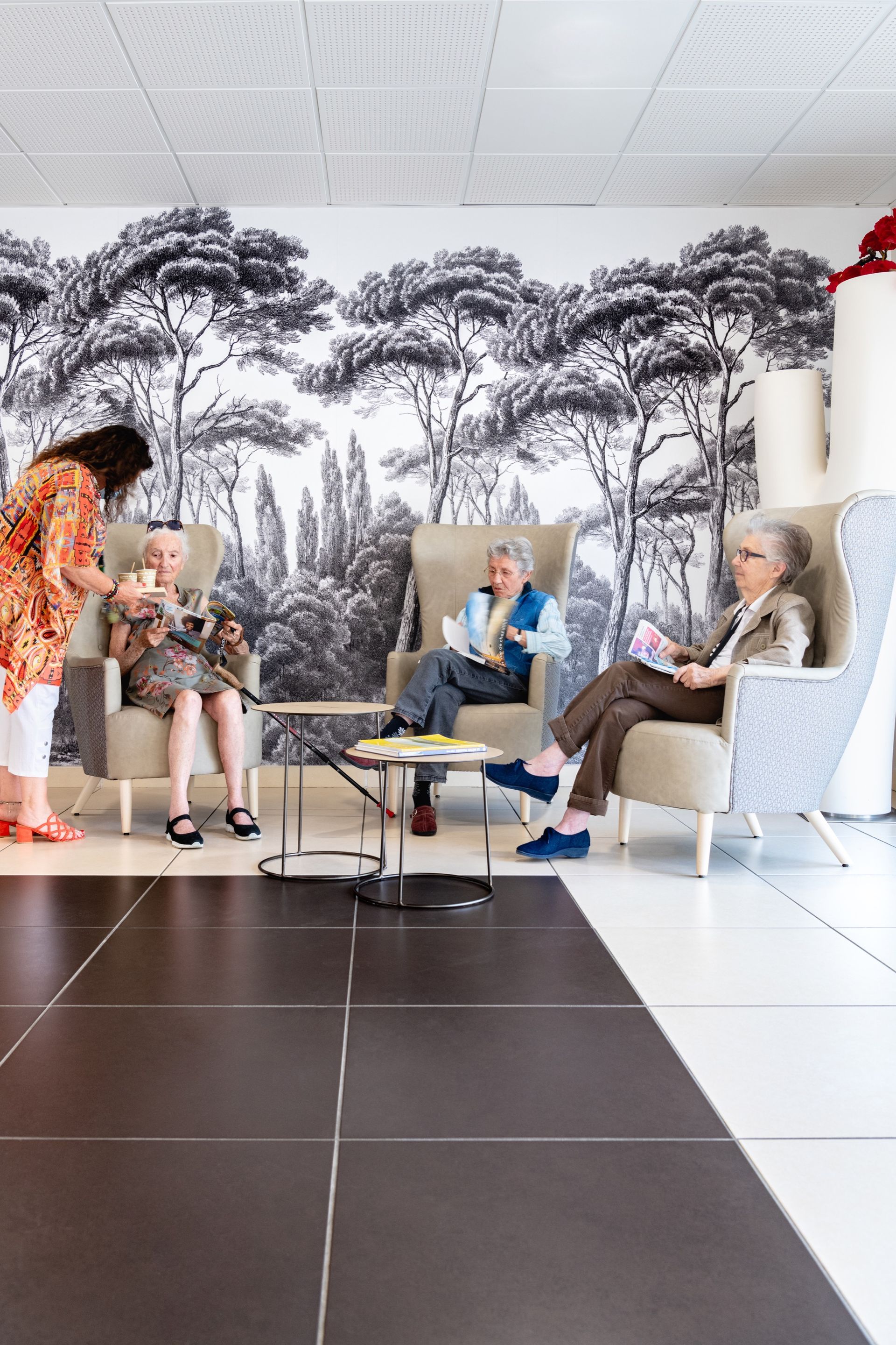 Quatre femmes âgées sont assises dans des fauteuils près d'une fresque murale en noir et blanc, tandis qu'une femme sert des rafraîchissements.