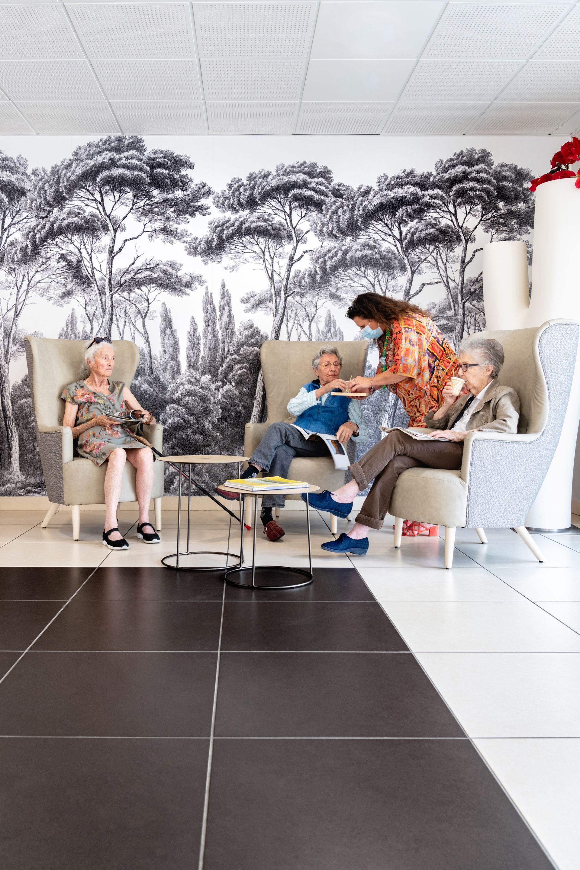 Quatre femmes âgées assises dans des fauteuils, l'une portant un masque, dans une pièce avec une fresque murale.