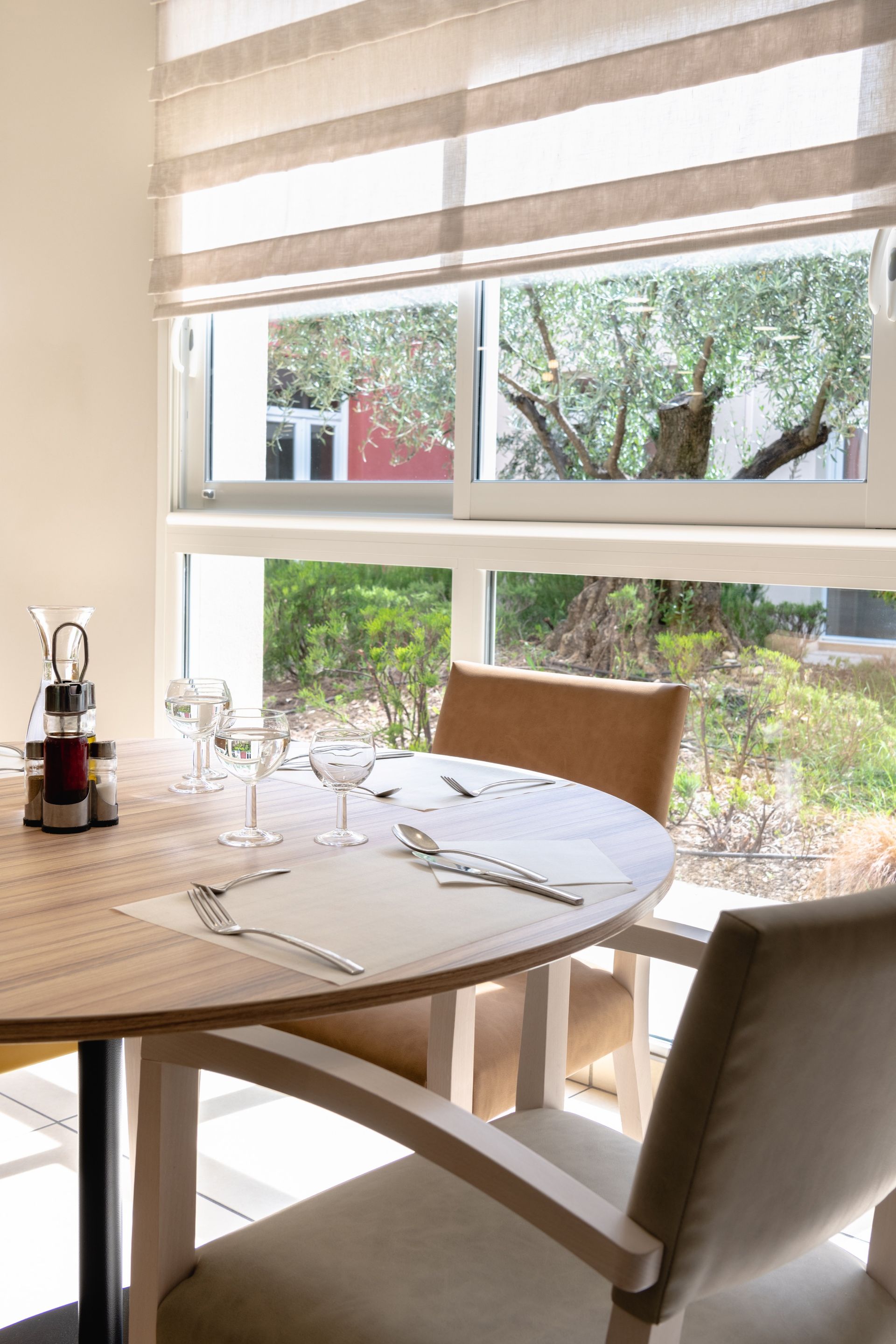 Ensemble table à manger ronde pour deux près d'une fenêtre donnant sur une cour verdoyante, deux chaises, tons beige clair et bois.