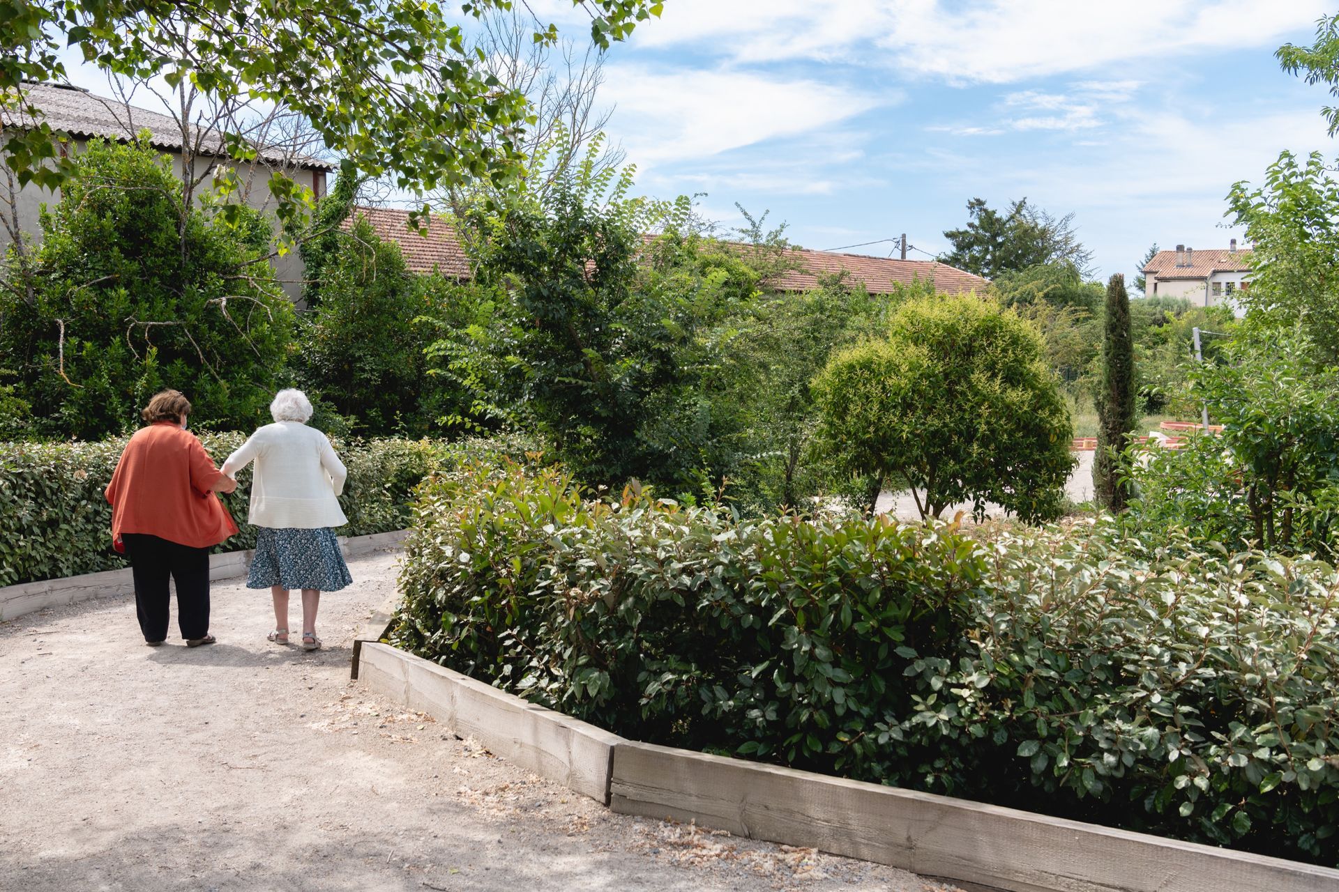 Deux femmes âgées marchent main dans la main le long d'un chemin de gravier entouré de buissons verts et d'arbres par une journée ensoleillée.