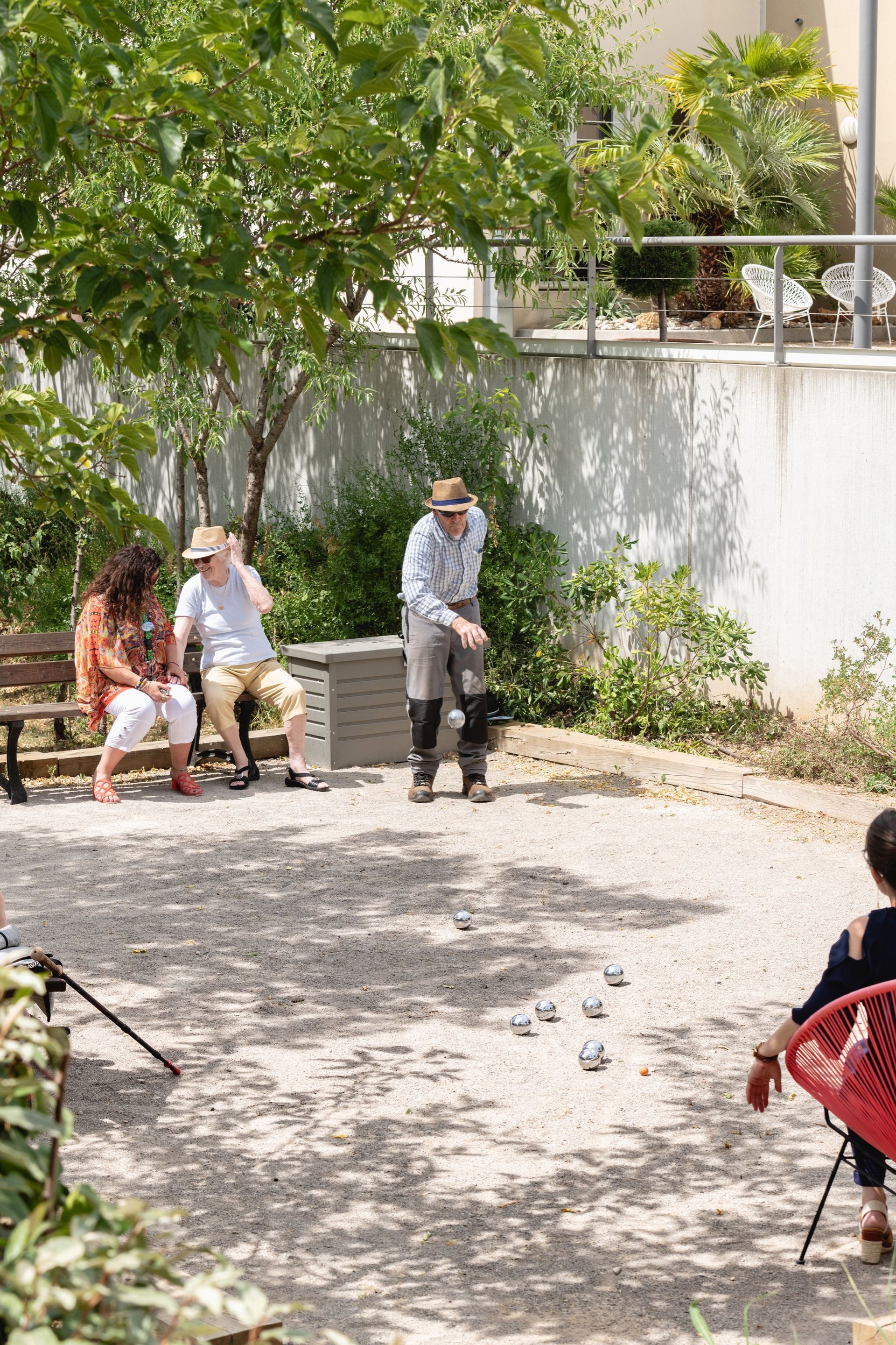 Des gens jouent à la pétanque sur un terrain en gravier. L'un lance une boule en métal ; d'autres regardent depuis des bancs et des chaises.
