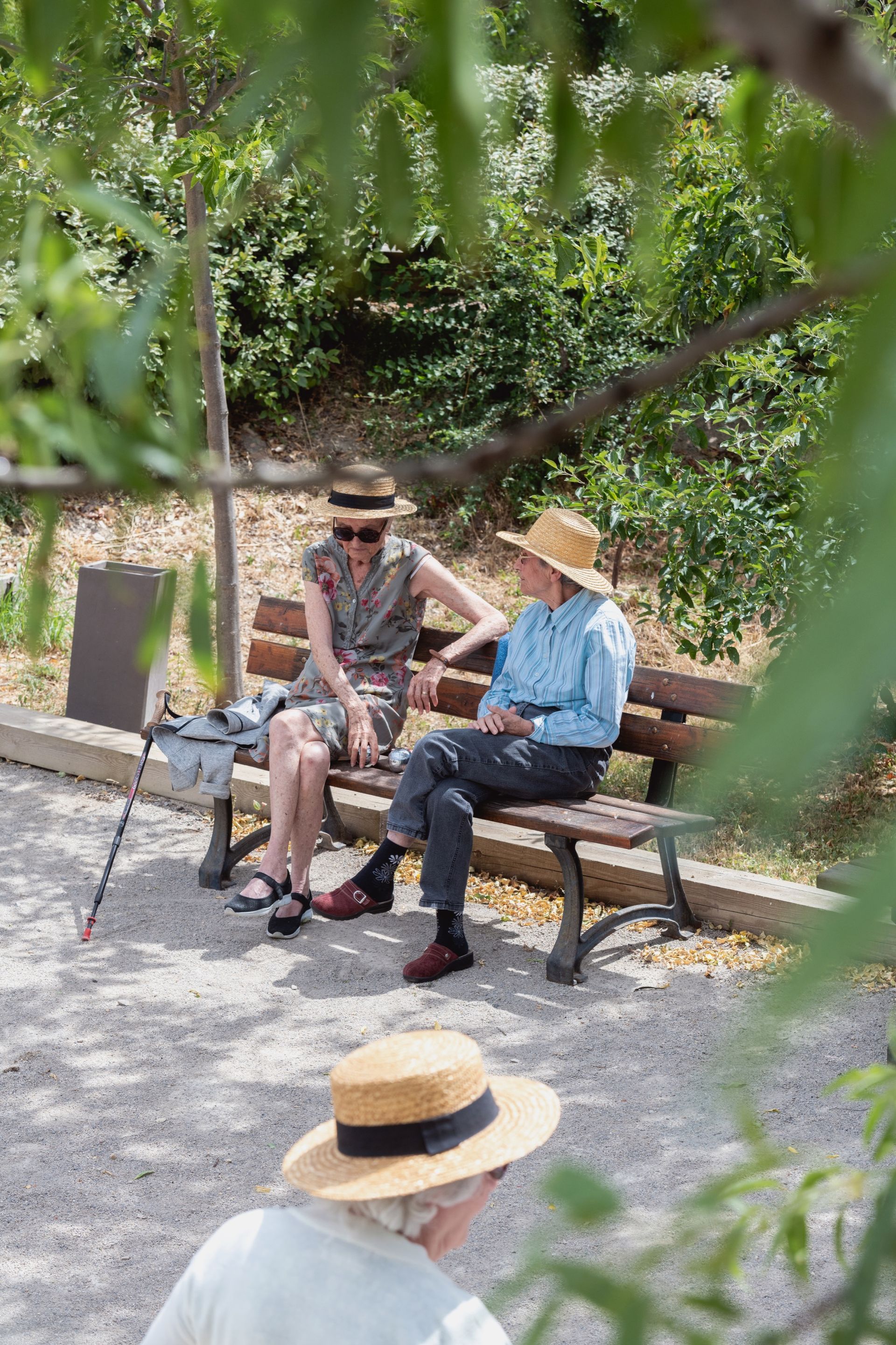 Deux personnes portant des chapeaux de paille sont assises sur un banc de parc et discutent ; une autre personne portant un chapeau joue à proximité.