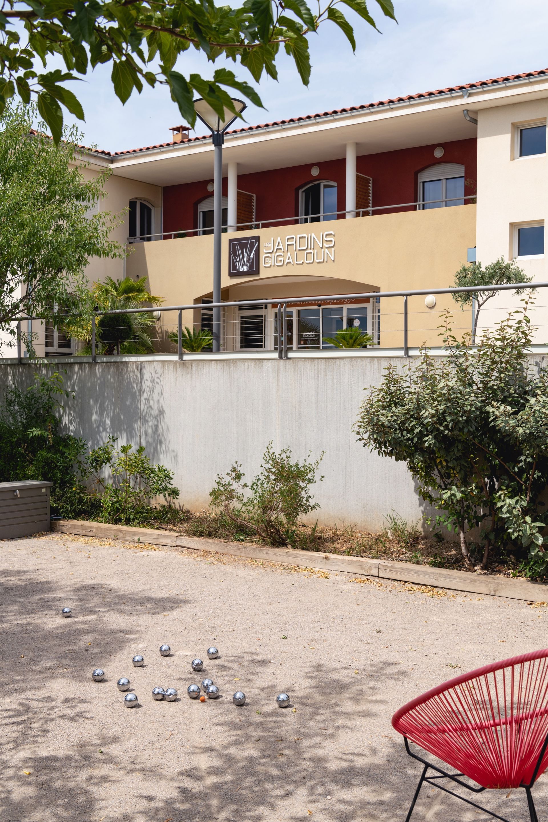 Un hôtel aux touches de rouge, avec un jeu de boules au premier plan. Une chaise rouge repose sur du gravier.