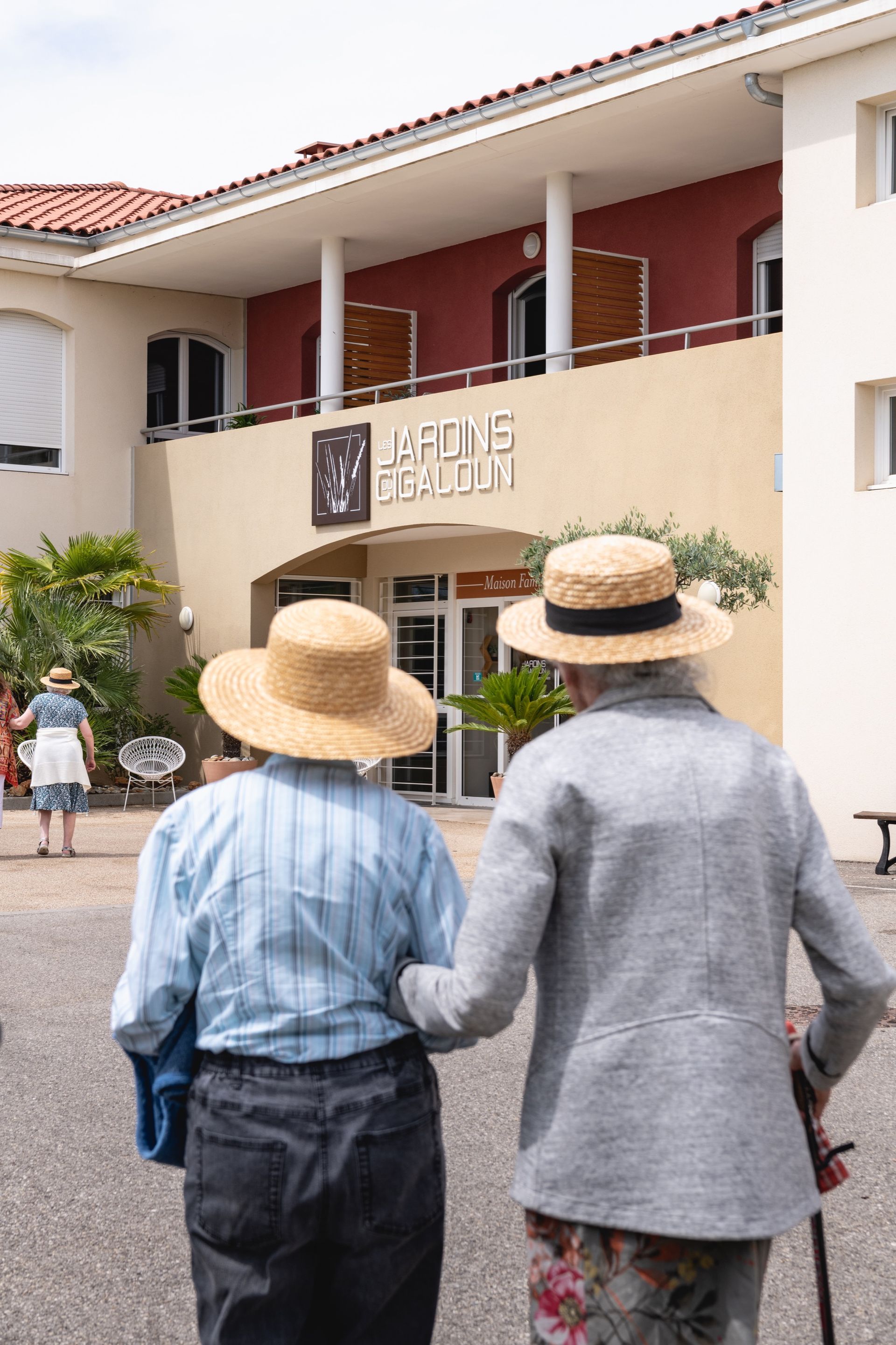 Deux femmes âgées portant des chapeaux de paille marchent bras dessus bras dessous vers la maison de retraite les Jardins du Cigaloun
