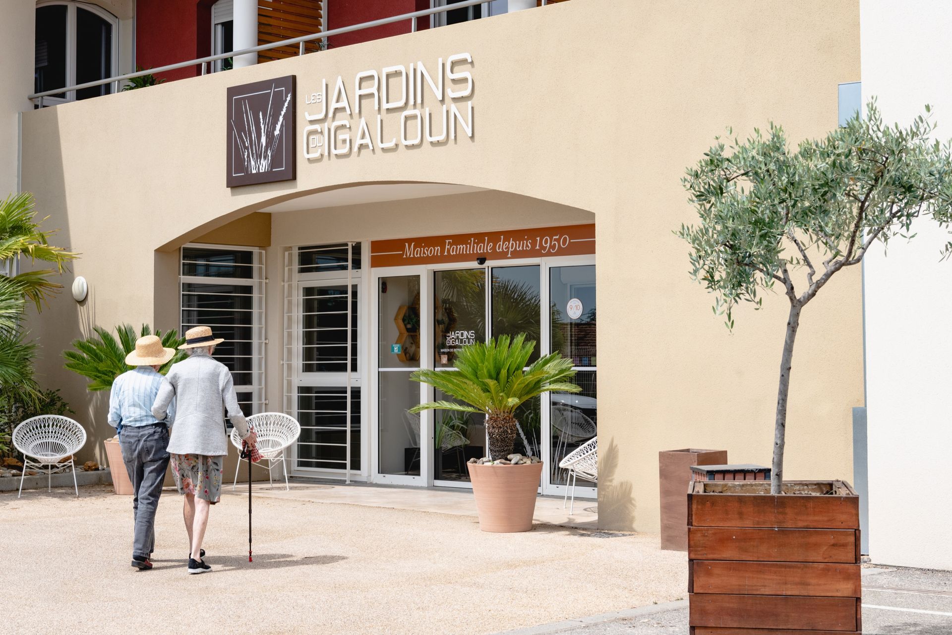 Deux personnes âgées se dirigent vers l'entrée des « Jardins Gigaloun ». Un bâtiment lumineux avec des portes vitrées, des palmiers et un olivier.