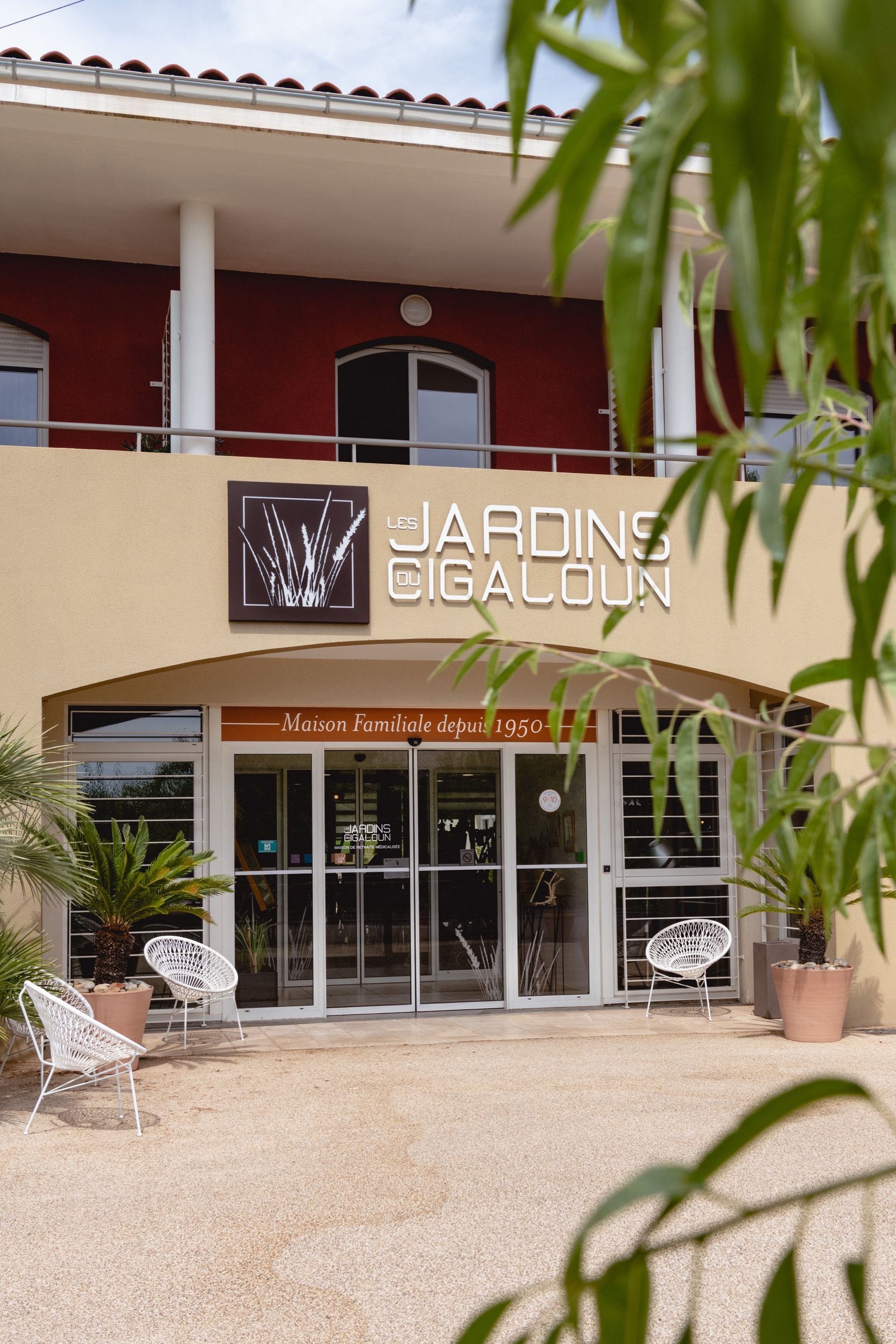Extérieur du bâtiment « Jardins Gigaloun » avec une façade beige, des portes vitrées et des sièges extérieurs.